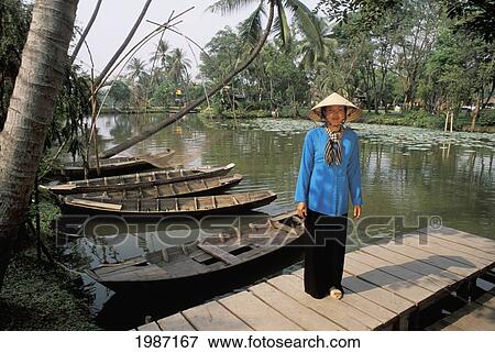 Stock Photo - Vietnam, Woman in traditional clothing standing on dock near river and old boats; Mekong Delta. Fotosearch