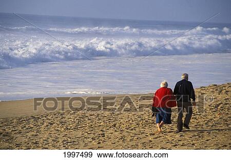 Stock Photo - View From Behind Senior Couple Walking On Beach, Arm In Arm White Shorebreak Ocean Water. Fotosearch
