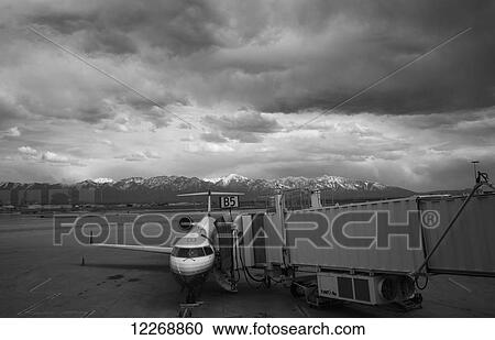 Stock Image - View of a commercial airline and the jet bridge with the Wasatch Mountains in the background on a cloudy overcast day, Salt Lake City International Airport, Utah. Fotosearch