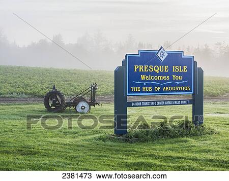 Stock Image - Welcome sign to Presque Isle, Maine on a foggy summer morning with farm equipment, a potato field and dewy grass surrounding it; Presque Isle, Maine, United States of America. Fotosearch