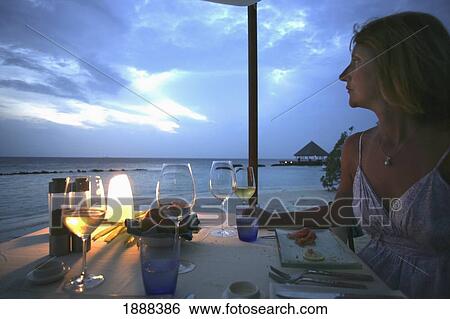 Stock Photograph - Woman At Dining Table Overlooking The Beach At Dusk.. Fotosearch