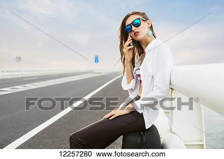 Stock Image - Woman calling with her mobile phone on the side of a highway, tired she sits in her luggage; Xiamen, Fujian, China. Fotosearch