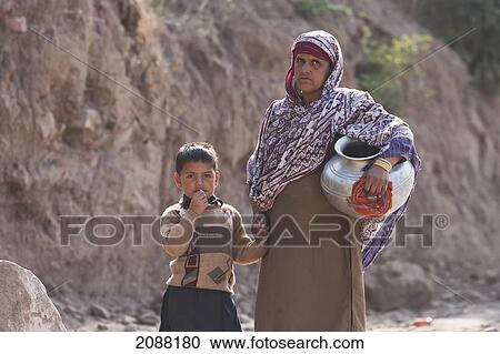 Stock Image - Woman Carrying A Water Jug Holding A Boy's Hand, Arja, Azad Kashmir, Pakistan. Fotosearch