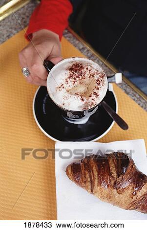 Stock Image - Woman In Cafe Holding A Cup Of Cappuccino With Croissant In Foreground. Fotosearch