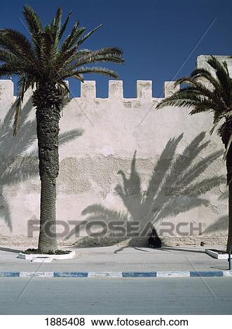 Stock Photo - Woman In Djelleba Resting In Shadow Of Date Palm Against Town Walls. Fotosearch