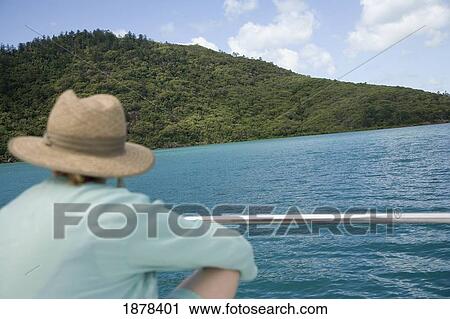 Stock Image - Woman In Hat Sitting On Deck Of Sail Boat Looking At Island. Fotosearch