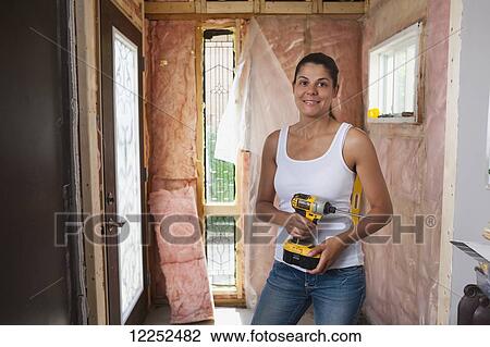 Stock Image - Woman in her 30s holding a drill at entrance of her home that is being renovated; Bradford, Ontario, Canada. Fotosearch