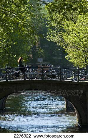 Stock Image - Woman on bicycle crossing bridge early in the morning; Amsterdam, Holland. Fotosearch