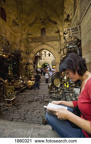 Stock Image - Woman Reading Guidebook Beside Stalls In Covered Gateway In Khan El Khalili. Fotosearch