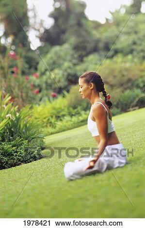 Stock Image - Woman sitting on grass doing yoga in tropical garden. Fotosearch