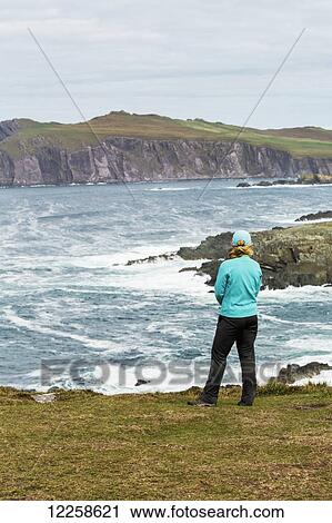 Stock Image - Woman standing on grassy cliff overlooking rugged shoreline with waves; Ballinglanna, County Cork, Ireland. Fotosearch