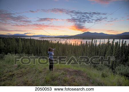 Stock Image - Woman Taking Photos At Sunset Of Teslin With Nisutlin Bay Bridge Over River, Yukon. Fotosearch