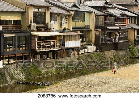 Stock Photograph - Woman Walking Along The River In Front Of Old Buildings; Kyoto, Japan. Fotosearch