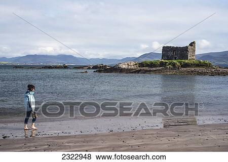 Stock Photo - Woman walking on beach with Ballinskelligs Castle in background; County Kerry, Ireland, UK. Fotosearch