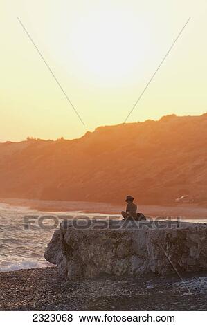 Stock Photo - Woman wearing bikini and sunhat, sitting on rock at sunset; Aphrodite Bay, Cyprus. Fotosearch
