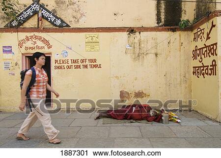 Stock Image - Woman With Backpack Walking Past Sadhu Sleeping On Pavement Outside Small Temple. Fotosearch