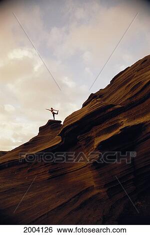 Stock Photograph - Woman Yoga At Sunrise On Rock Coastline Cliff, Side View Upward Against Clouds. Fotosearch