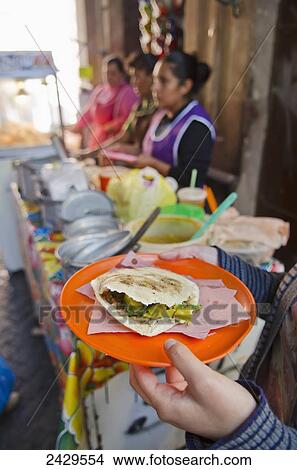 Picture - Woman's hands holding a plate of indigenous mexican food, a gordita, beside the food vendor stand; San Luis de la Paz, Guanajuato, Mexico. Fotosearch