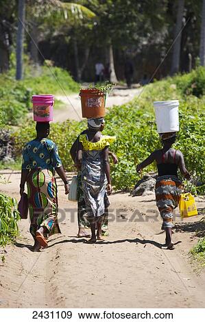Stock Photo - Women balancing large buckets on their heads as they walk down the dirt road; Vamizi Island, Mozambique. Fotosearch
