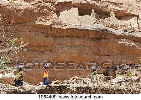 Stock Photo - Women carrying container on head near Telem houses set in the Bandiagara Escarpment, Irelli, Mopti, Mali. Fotosearch