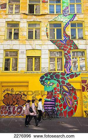 Stock Photograph - Women walking past large mural of snake and businessman painted on front of building; Amsterdam, Holland. Fotosearch