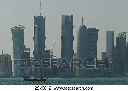 Stock Image - Wooden boat crossing harbour in front of the modern skyline of Doha; Doha, Qatar. Fotosearch