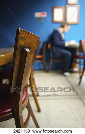 Stock Photo - Wooden chairs up against a wooden table in a cafe with a man sitting and reading at a table in the background; London, England. Fotosearch