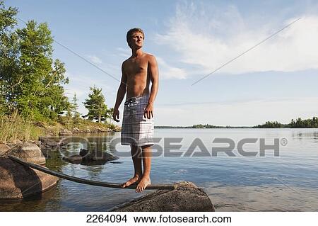 Picture - Young Adult Man Balancing On A Stick By A Rocky Lake Shoreline; Kenora Ontario Canada. Fotosearch