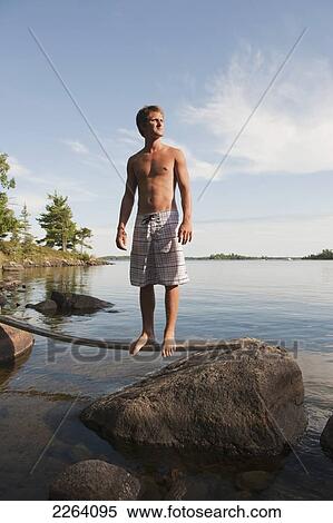 Stock Photography - Young Adult Man Balancing On A Stick By A Rocky Lake Shoreline; Kenora Ontario Canada. Fotosearch