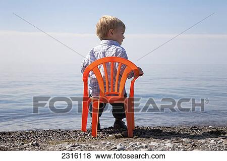 Stock Photo - Young Boy Sitting On An Orange Chair By The Shore Of Lake Ontario; Ontario Canada. Fotosearch