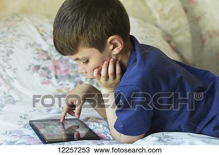 Stock Image - Young boy using a computer tablet on a bed; Lemington, Newcastle Upon Tyne, England. Fotosearch