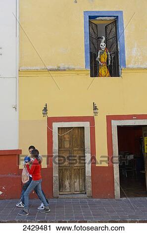 Stock Image - Young couple walking beneath a skeleton statue on an old Mexican street; Guanajuato, Mexico. Fotosearch