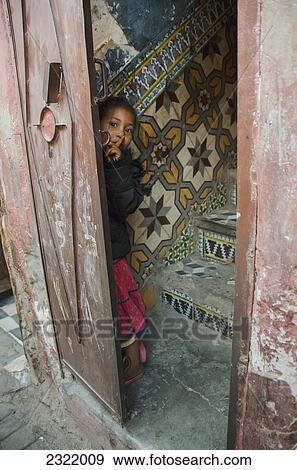 Stock Photo - Young girl hiding behind doorway with her finger to her lips indicating silence; Medina, Marrakech, Morocco. Fotosearch