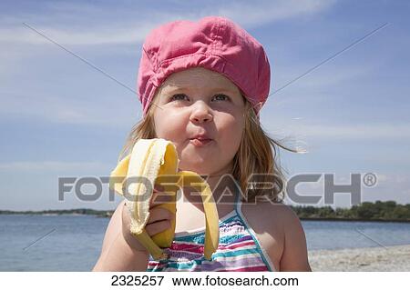 Stock Photo - Young girl in a pink hat enjoying a banana at the beach; Ontario canada. Fotosearch