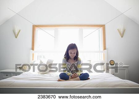 Stock Image - Young girl sitting cross-legged on her bed; Victoria, Vancouver Island, British Columbia, Canada. Fotosearch