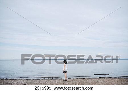 Stock Photography - Young Girl Standing On A Beach By The Water; Saint-Simeon Quebec Canada. Fotosearch