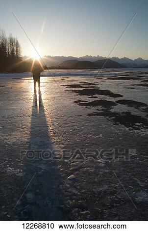 Stock Image - Young man standing on a frozen lake with arms raised and sunburst over his head; Haines, Alaska, United States of America. Fotosearch