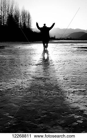 Stock Image - Young man standing on a frozen lake with arms raised; Haines, Alaska, United States of America. Fotosearch