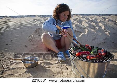 Stock Image - Young woman barbecuing seafood on the beach.. Fotosearch
