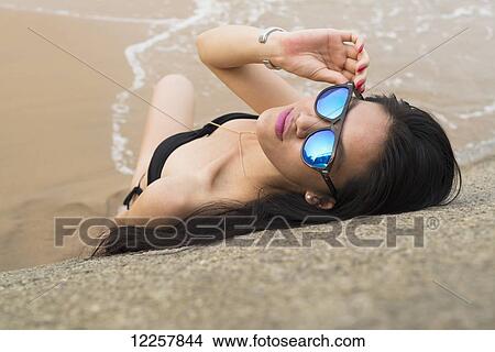 Picture - Young woman laying on the beach at the water's edge in a bikini and sunglasses; Xiamen, China. Fotosearch