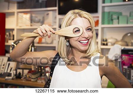 Stock Photography - Young woman looking through a hole in a mixing spoon at a kitchen store in shopping mall; St. Albert, Alberta, Canada. Fotosearch