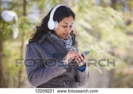 Stock Image - Young woman of South Asian ethnicity using cell phone and Bluetooth headphones in Scanlon Creek Conservation Area; Ontario, Canada. Fotosearch