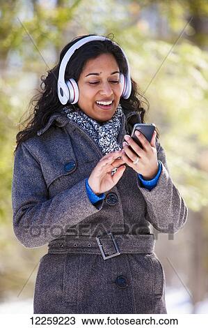 Stock Image - Young woman of South Asian ethnicity using cell phone and Bluetooth headphones in Scanlon Creek Conservation Area; Ontario, Canada. Fotosearch