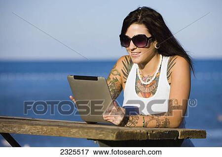Stock Photo - Young woman sitting at a picnic table by a lake using a tablet; Ontario canada. Fotosearch