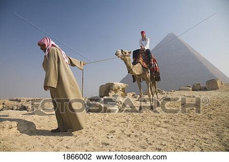 Stock Image - Young Woman Tourist Riding A Camel Lead By A Guide At The Pyramids Of Giza, Egypt. Fotosearch