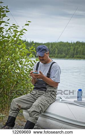 Stock Photo - A Alaskan Native man using a smart phone while sitting on a rubber boat along the Kenai River shore, Kenai Wildlife Refuge, Southcentral Alaska, Summer. Fotosearch