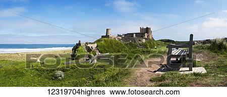 Stock Photograph - A bench with a view of the ocean and Bamburgh Castle in the distance; Bamburgh, Northumberland, England. Fotosearch