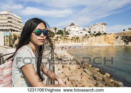 Stock Photograph - A Chinese young woman in sunglasses stands at a railing looking out at the water; Benidorm, Alicante, Spain. Fotosearch
