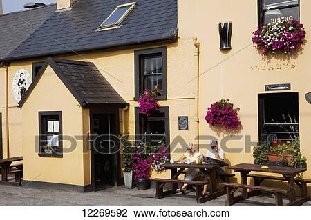 Stock Image - A couple sits at a table in front of a pub in a yellow building; Caherdaniel, County Kerry, Ireland. Fotosearch
