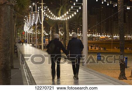 Stock Image - A couple walking and holding hands along a promenade on the beach; Benidorm, Spain. Fotosearch